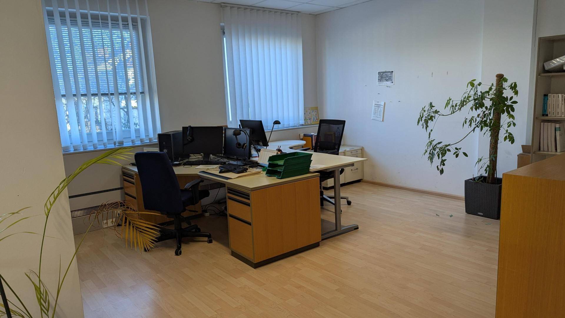 Helles Büro mit mehreren Arbeitsplätzen, hellem Laminatboden und vertikalen Jalousien an den Fenstern.