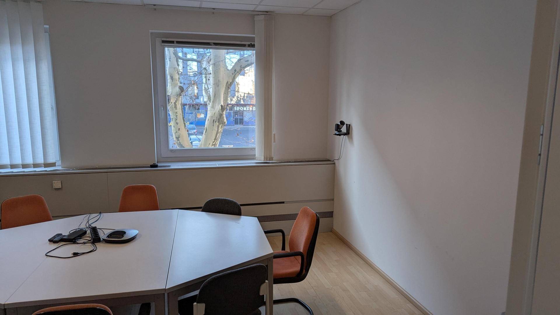 Schlichter Konferenzraum mit einem achteckigen Tisch, Stühlen und einem Fenster mit Blick nach draußen.