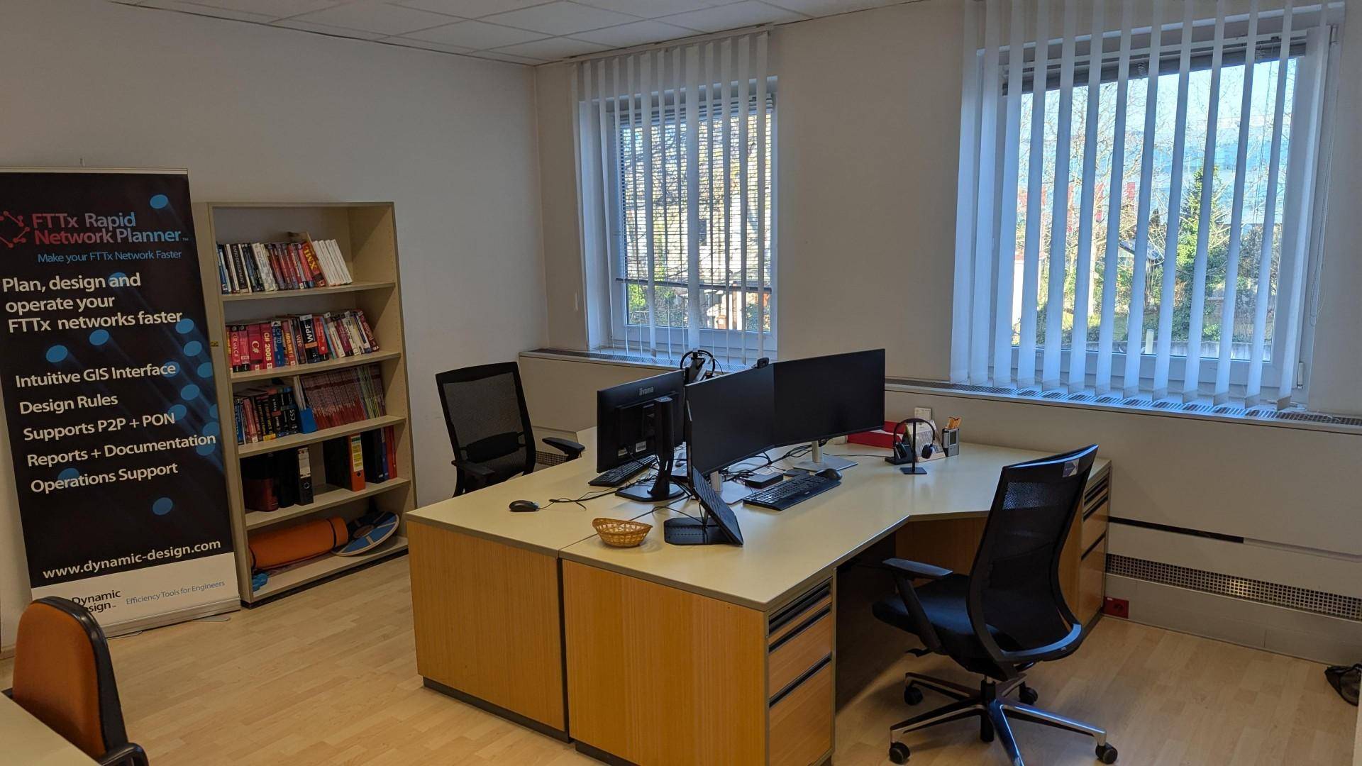Modern eingerichtetes Büro mit Schreibtischen, Bürostühlen und einem Bücherregal neben den Fenstern.