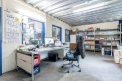 Büroarbeitsplatz in einem Lagerraum mit Schreibtisch, Bürostuhl und Regalen im Hintergrund.