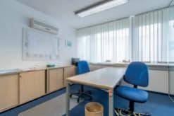 Helles Büro mit Schreibtisch, blauen Bürostühlen und Fenster mit vertikalen Jalousien.