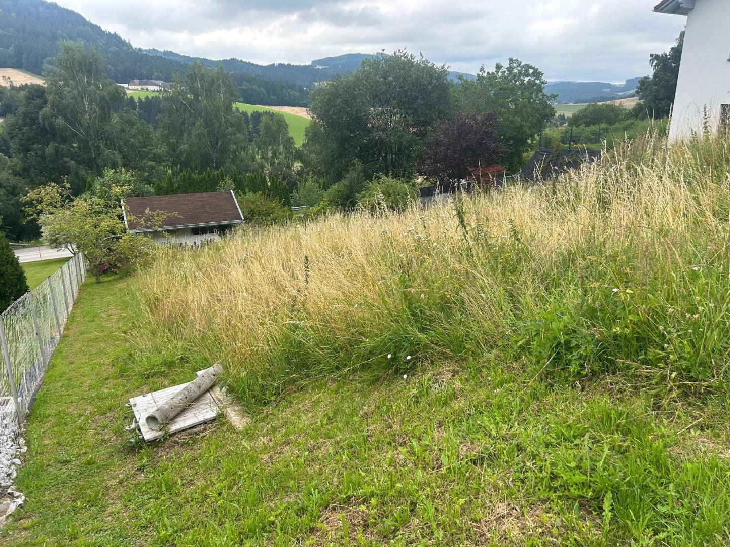 Blick über ein grasbewachsenes Grundstück mit einem Haus und grünen Hügeln im Hintergrund.
