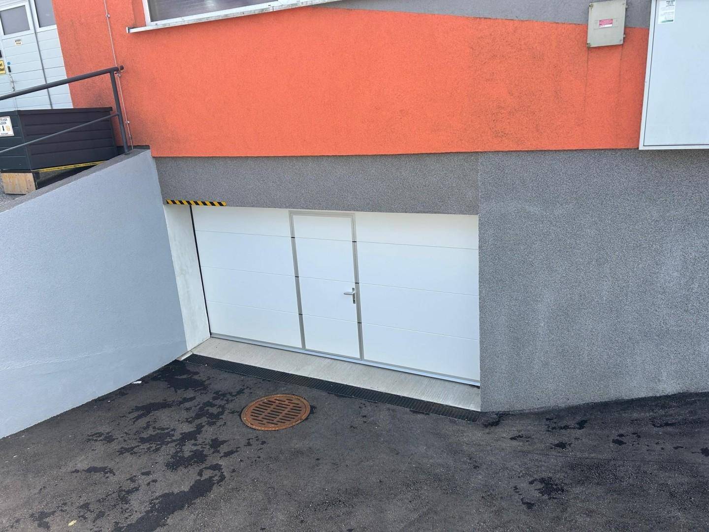 Nahaufnahme der weißen Tiefgarageneinfahrt mit asphaltierter Zufahrt und Entwässerungsrinne am Boden.