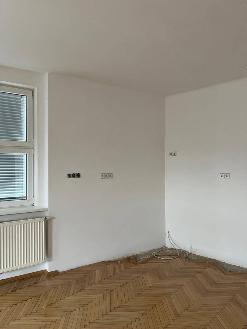 Leere Raumecke mit Fischgrätparkett, Fenster und Heizkörper, bereit für die Einrichtung.