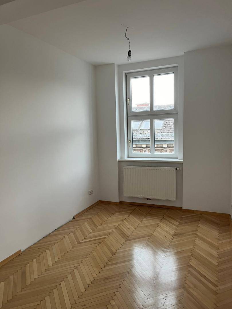 Helles, leeres Zimmer mit Fischgrätparkett und einem großen Fenster, ideal als Schlafzimmer.