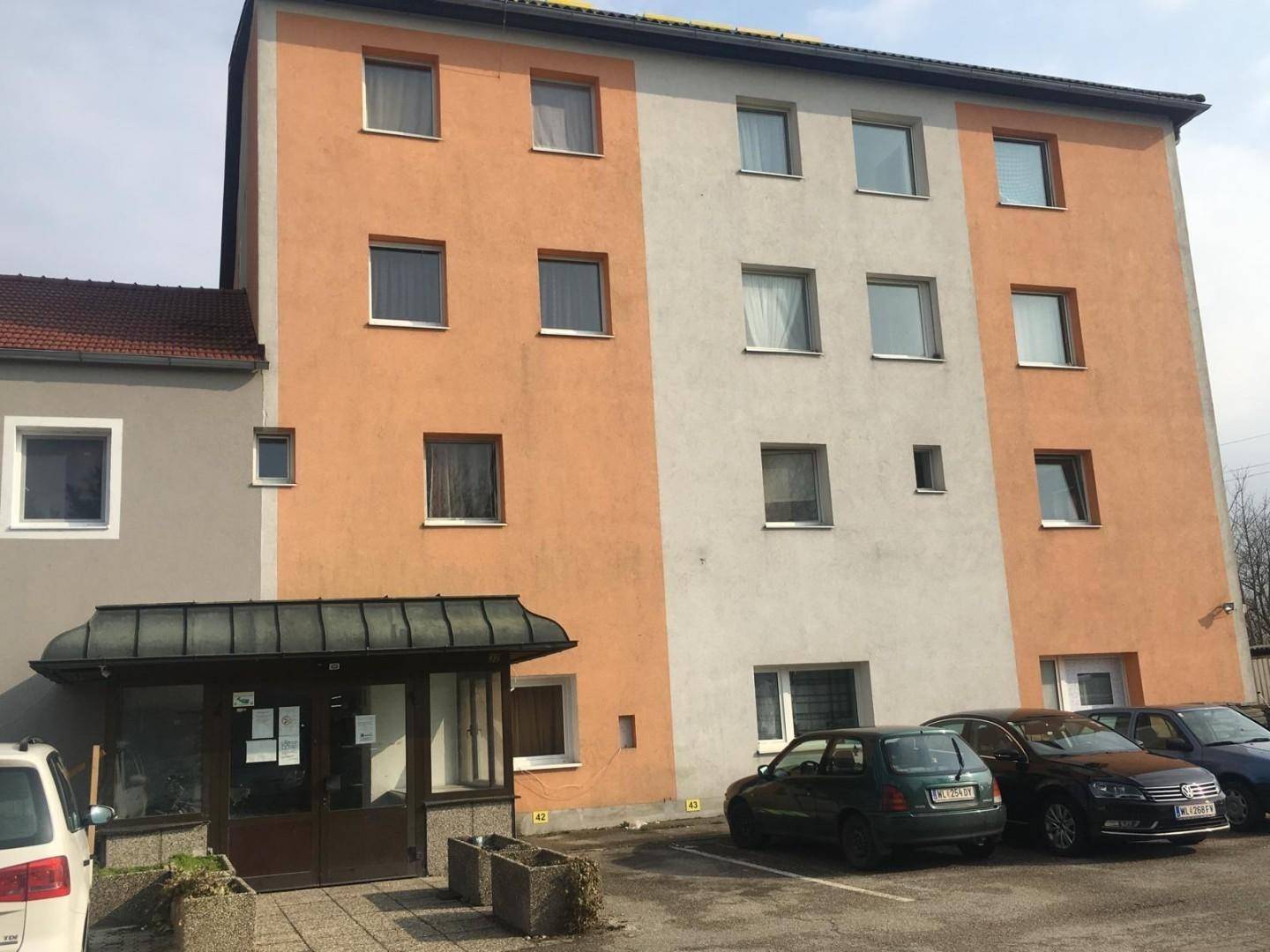 Vorderansicht eines mehrstöckigen Gebäudes mit orange-weißer Fassade, Eingangsbereich und geparkten Autos.