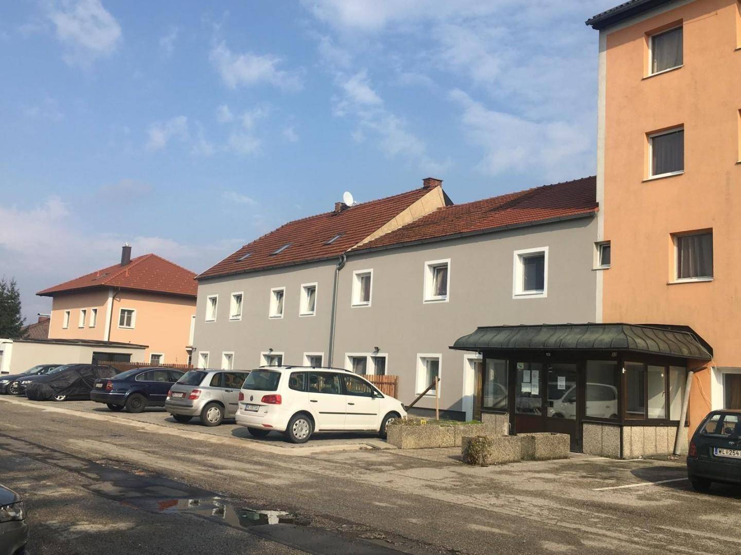 Außenansicht eines grauen Gebäudes mit braunem Dach neben einem orangefarbenen Gebäude und Parkplätzen.