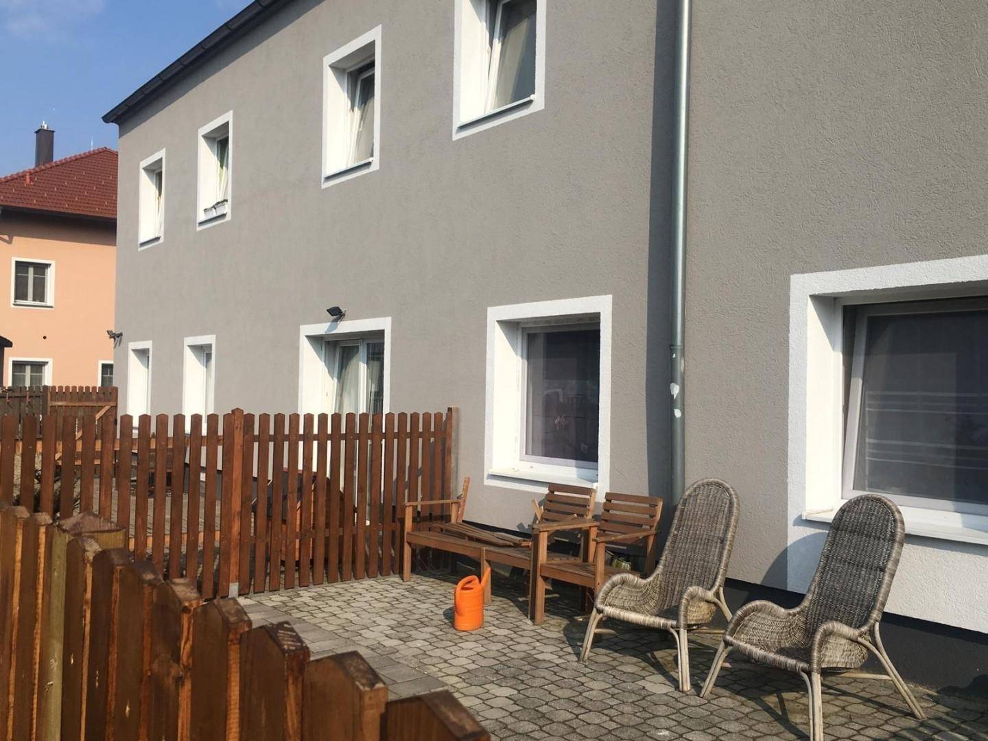 Gepflasterte Terrasse neben einem grauen Gebäude mit Holzzaun, Holzbank und Korbstühlen.