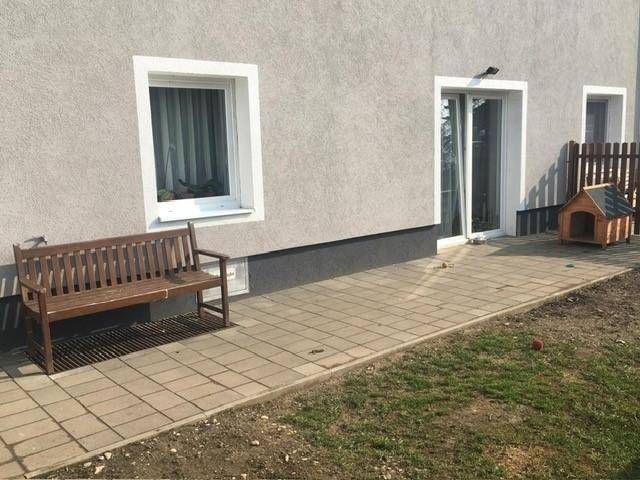 Gepflasterter Bereich neben einem grauen Gebäude mit Holzbank, Glasschiebetür und Rasenfläche.