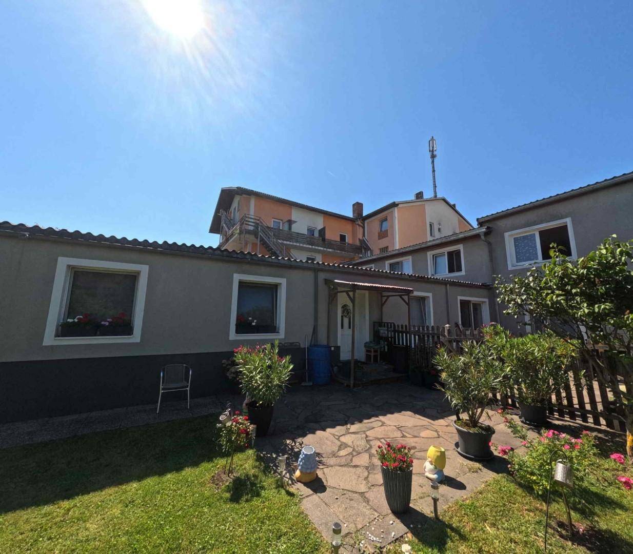 Sonniger Hinterhof mit gepflasterter Terrasse, Rasenfläche und Topfpflanzen vor einem grauen Gebäude.