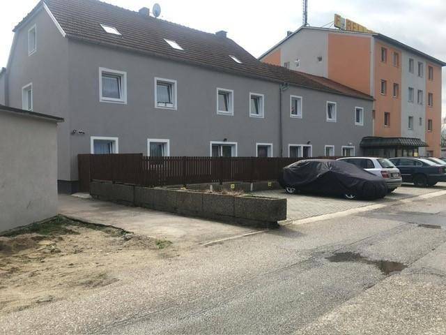 Außenansicht eines grauen Gebäudes mit braunem Dach, Holzzaun und geparkten Autos auf dem Gelände.