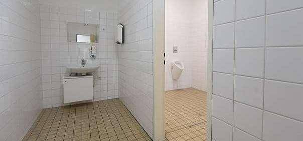 Gefliester Toilettenbereich mit Waschbecken, Spiegel und Urinal in einem angrenzenden Raum.