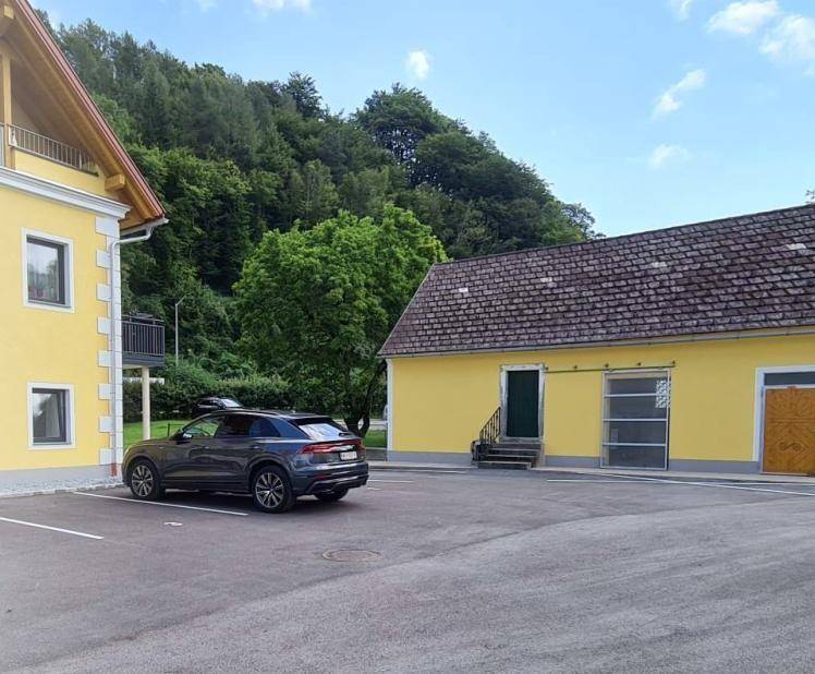 Gelbe Gebäude mit Parkplätzen und einem Auto, umgeben von grünen Bäumen und Hügeln.