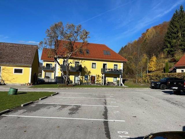 Gelbes Wohngebäude mit roten Ziegeldächern und Balkonen, umgeben von Natur und Parkplätzen.