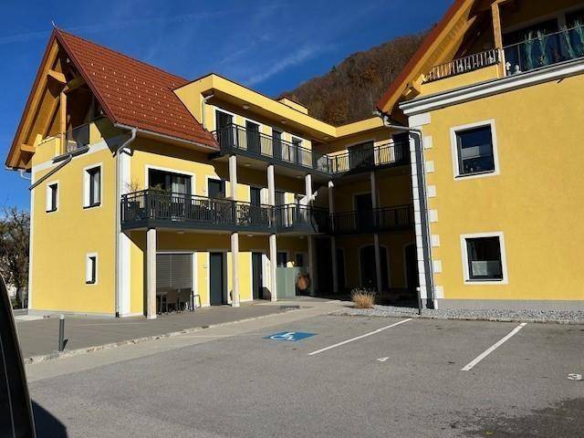 Gelbes Mehrfamilienhaus mit mehreren Balkonen und einem barrierefreien Parkplatz im Vordergrund.
