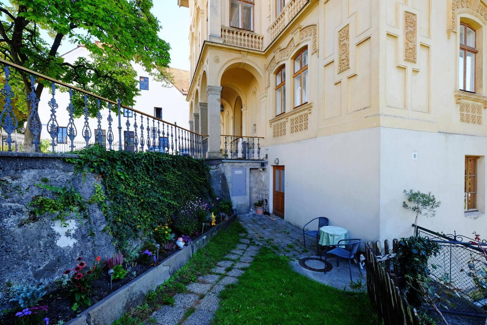 Kleiner Gartenbereich mit gepflastertem Weg, Blumenbeeten und Sitzgelegenheit im Freien.