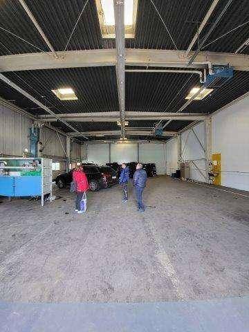 Geräumige Lagerhalle mit hoher Decke, Oberlichtern und sichtbaren Stahlträgern, in der sich Personen und Fahrzeuge befinden.