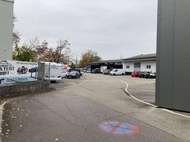 Weitläufiger Außenbereich eines Gewerbeparks mit Parkplätzen, Bäumen und weiteren Gebäuden im Hintergrund.