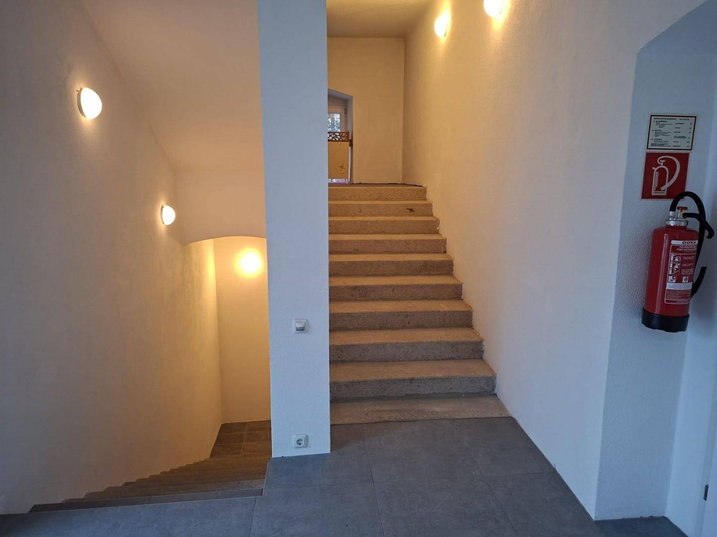 Treppenhaus mit Steinstufen, weißen Wänden und Wandleuchten, führt zu oberen und unteren Etagen.