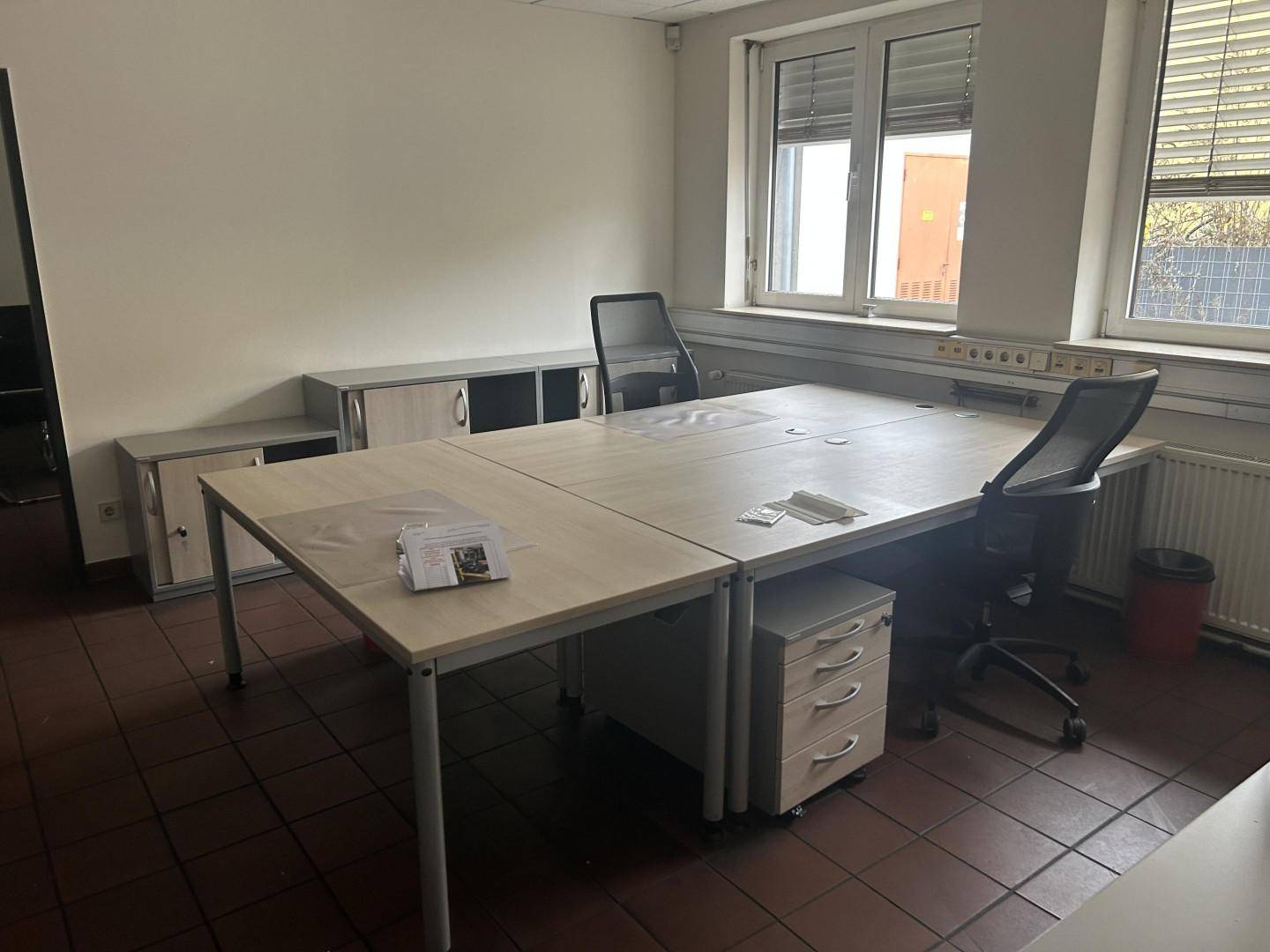Funktionales Büro mit mehreren Arbeitsplätzen, Rollcontainern und Fliesenboden, gut beleuchtet durch große Fenster.