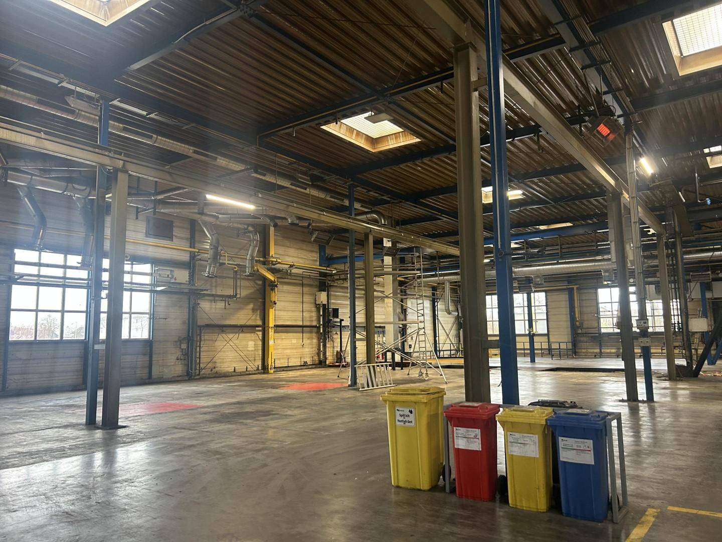 Weitläufige Industriehalle mit hohen Decken, großen Fenstern und Mülltonnen zur Abfalltrennung, mit viel Tageslicht.