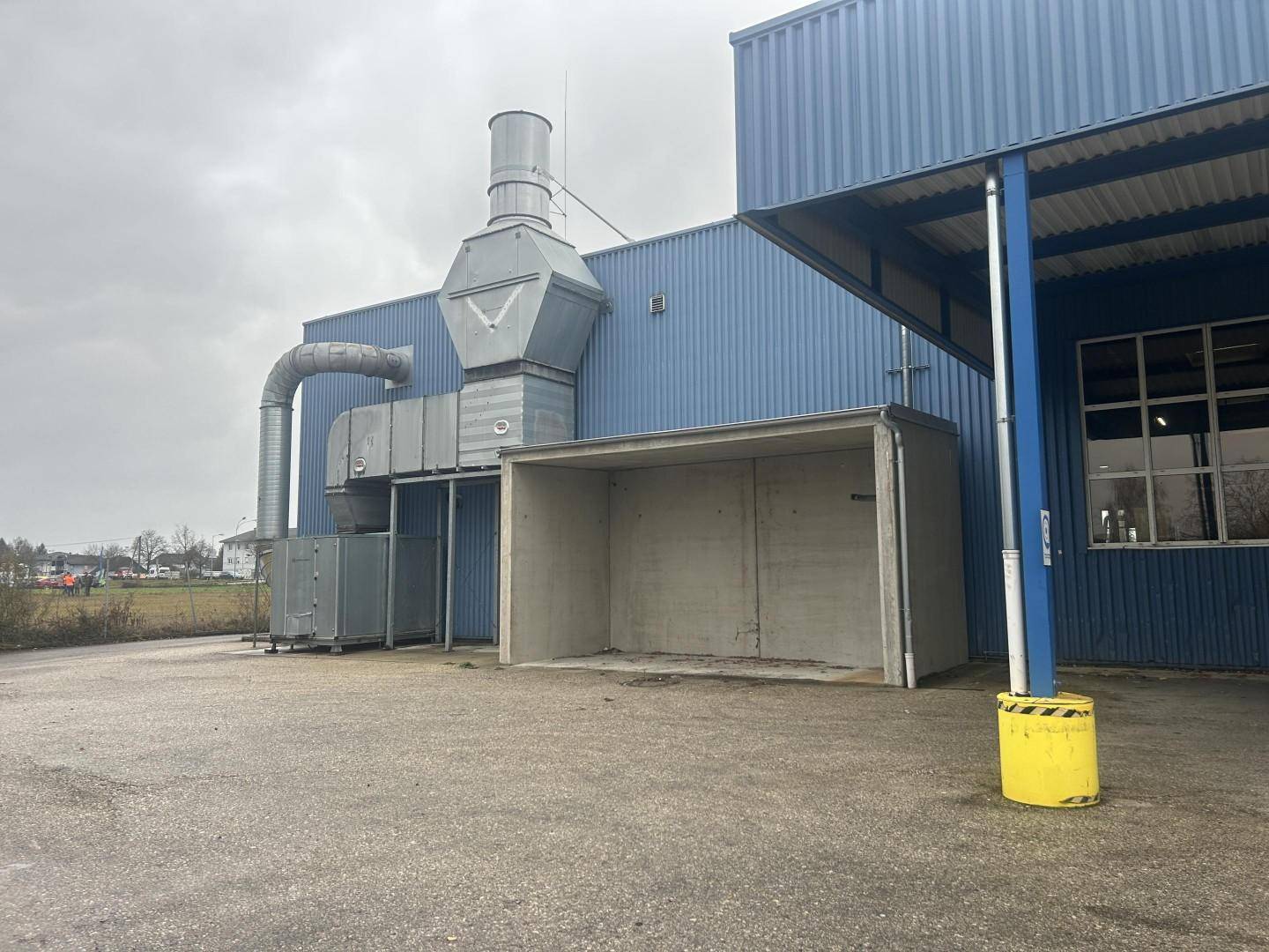 Industrielles Gebäude mit großen Lüftungsrohren und einem überdachten Bereich an der Seite, umgeben von einem asphaltierten Parkplatz.