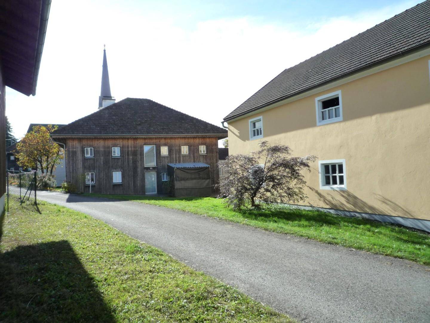 Ländliche Ansicht mit zwei Gebäuden, einer Zufahrt und einem Kirchturm in der Ferne.