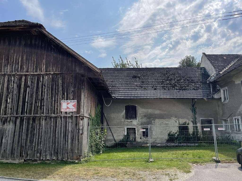 Altes, verwittertes Gebäude mit angrenzender Holzscheune und provisorischem Zaun im Vordergrund.