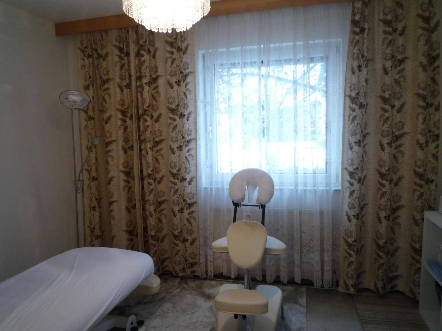 Raum mit Massageliege, Stuhl und einem Fenster mit gemusterten Vorhängen.