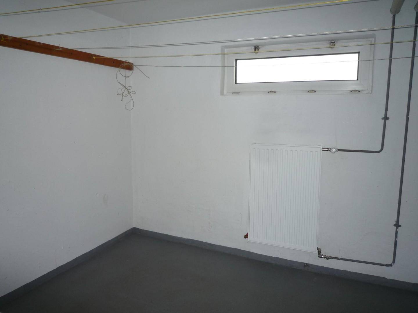 Leerer Kellerraum mit Fenster, Heizkörper und sichtbaren Rohrleitungen an der Wand.