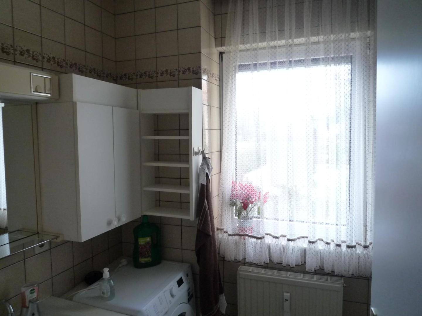 Badezimmer mit Fenster, Waschmaschine, Waschbecken und weißen Hängeschränken.
