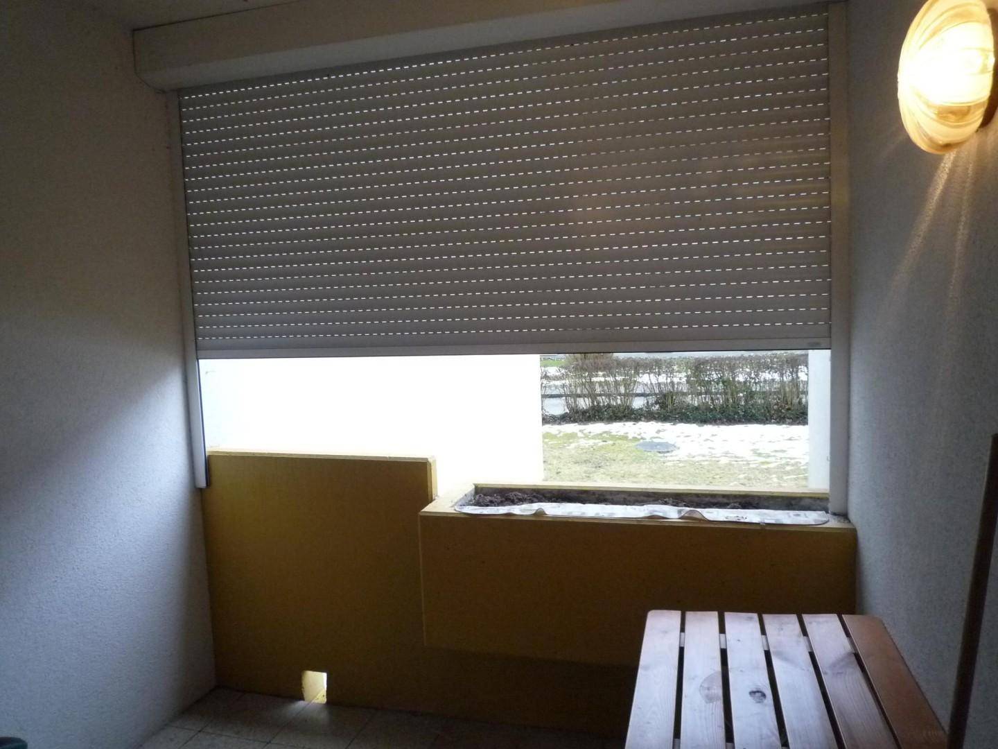Balkon mit heruntergelassenem Rollladen, kleinem Holztisch und Blick auf die verschneite Umgebung.