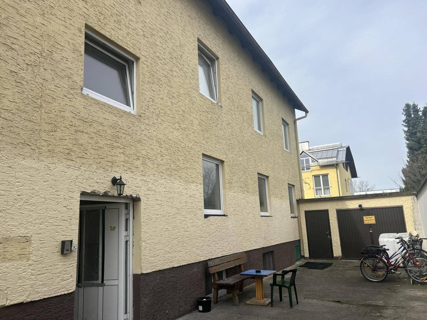 Seitenansicht des Gebäudes mit Eingangsbereich, Garagen und einem kleinen Sitzbereich im Freien.