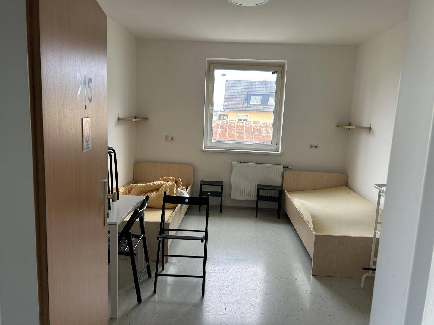 Einfaches Schlafzimmer mit zwei Einzelbetten, einem Fenster und funktionaler Einrichtung.