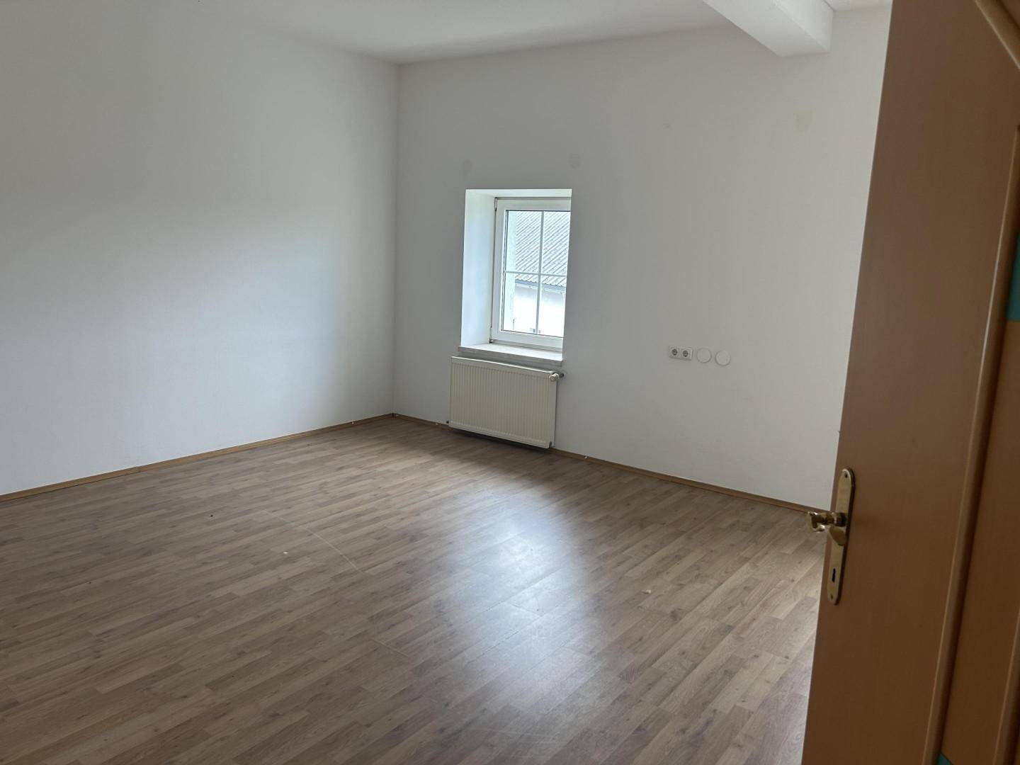 Leeres Zimmer mit Laminatboden, weißen Wänden und einem Fenster mit Blick nach draußen.
