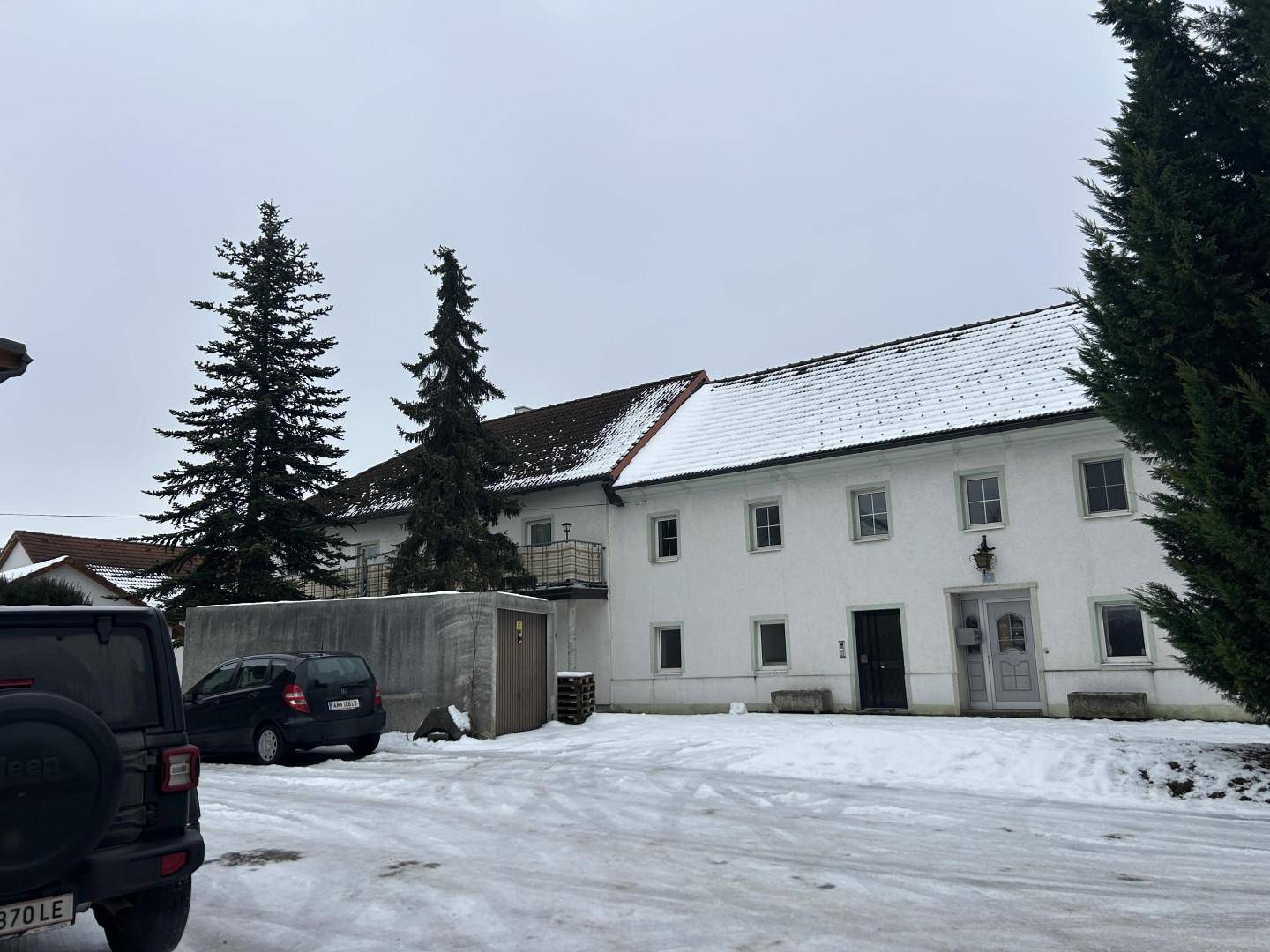 Vorderansicht eines weißen Gebäudes mit geparkten Autos und hohen Bäumen im verschneiten Winter.