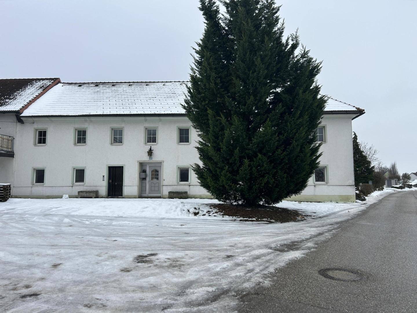 Vorderansicht eines langen weißen Gebäudes mit hohen Bäumen und verschneiter Umgebung.