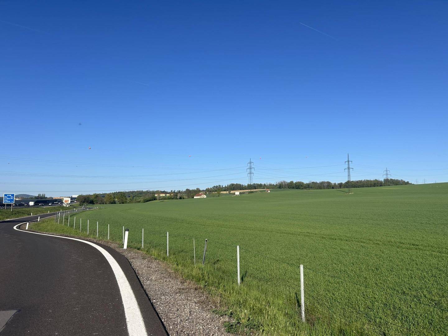 Panoramablick über weite grüne Felder mit einer Straße im Vordergrund und Industriegebäuden am Horizont.