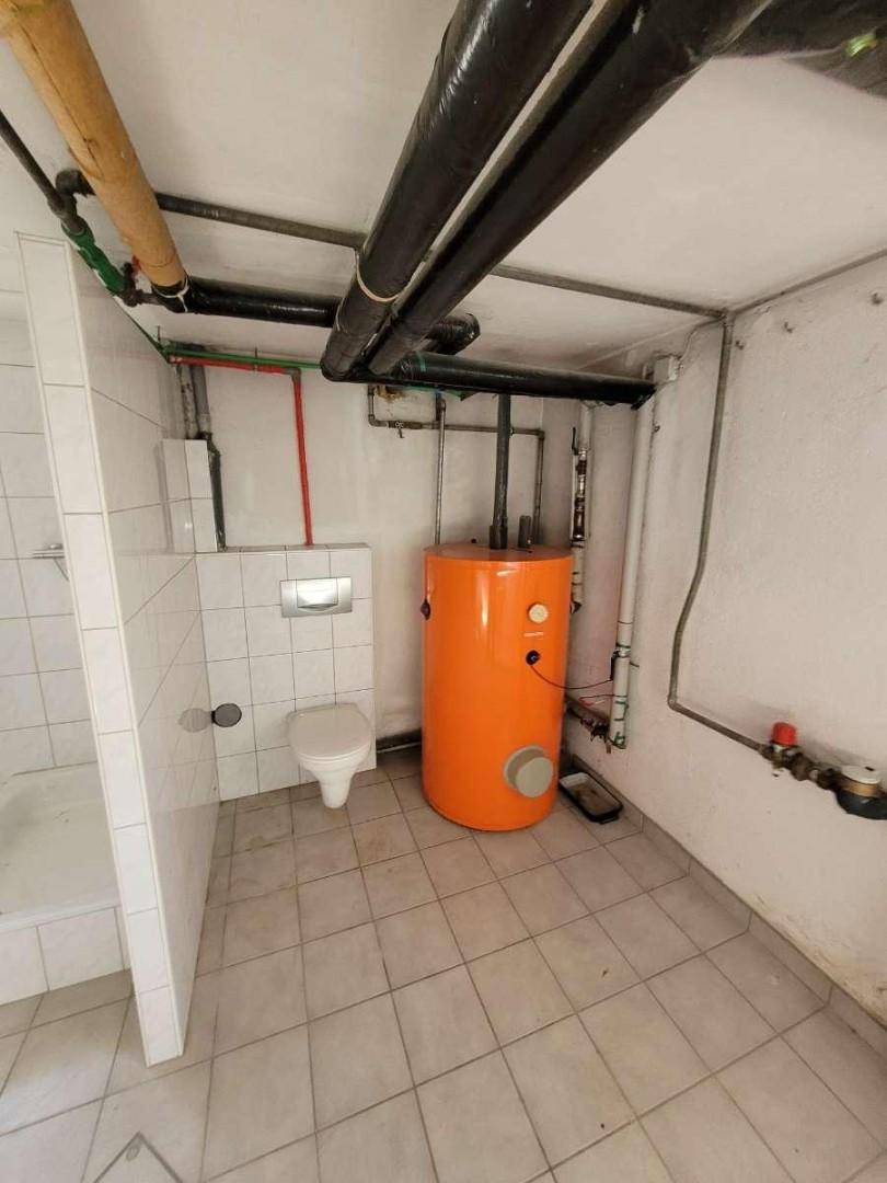 Funktionaler Kellerraum mit Boiler, Toilette und sichtbaren Rohrleitungen an der Decke.