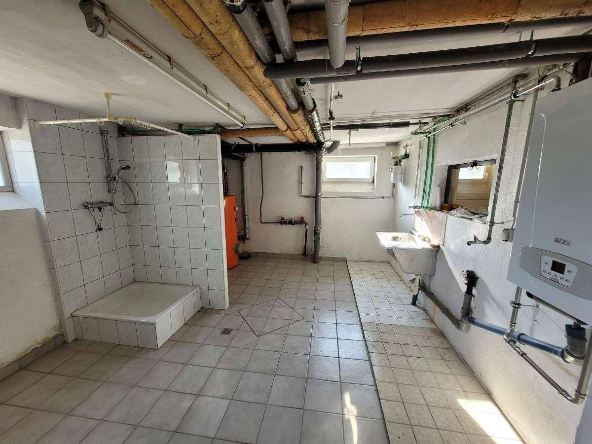Kellerraum mit Fliesenboden, sichtbaren Rohren, Dusche und Heizungsanlage.