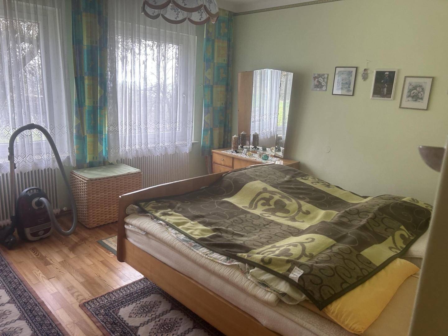 Schlafzimmer mit Holzboden, Doppelbett und zwei Fenstern mit Vorhängen.