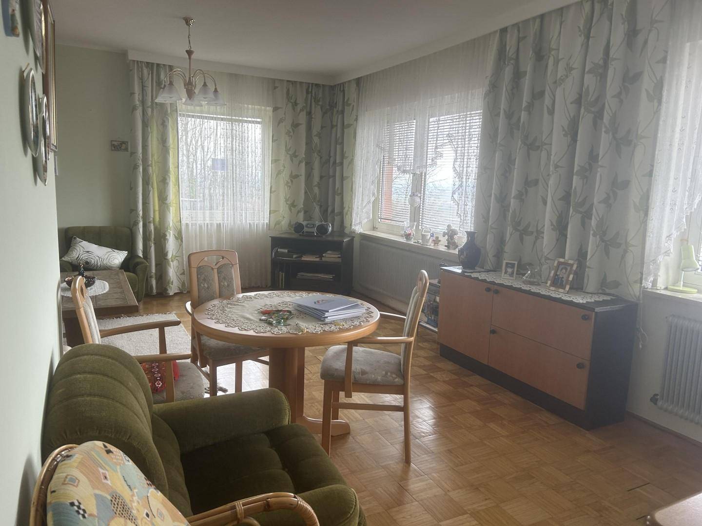 Helles Wohnzimmer mit Parkettboden, bequemen Sesseln und großen Fenstern mit Vorhängen.