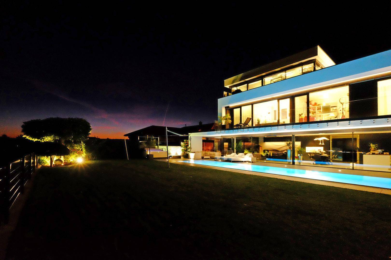 Elegante moderne Villa bei Nacht mit beleuchtetem Pool und warmen Innenlichtern.