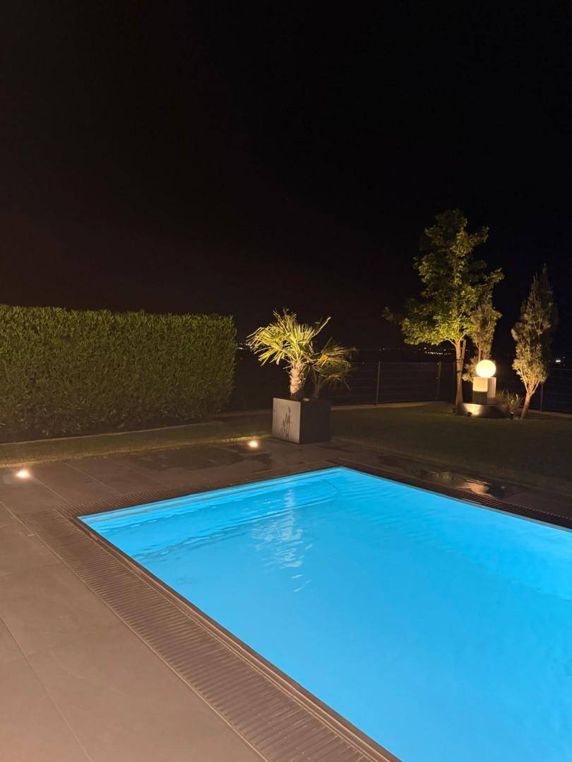 Beleuchteter Poolbereich im Garten bei Nacht, umgeben von Hecken und Bäumen.