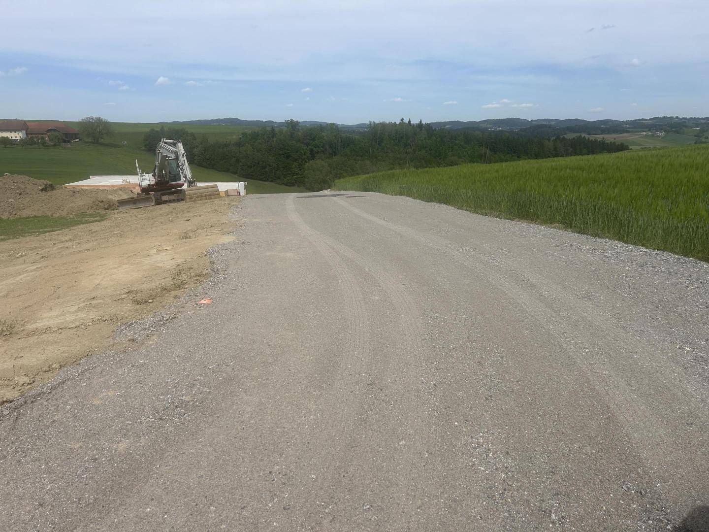 Schotterweg führt durch grüne Felder, mit einem Bagger im Hintergrund, bereit für Bauarbeiten.