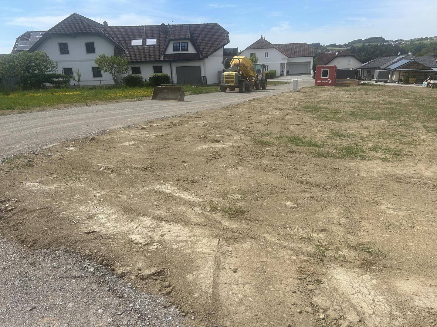 Baustelle mit Schotterfläche und einem Zementmischer, umgeben von Wohnhäusern.