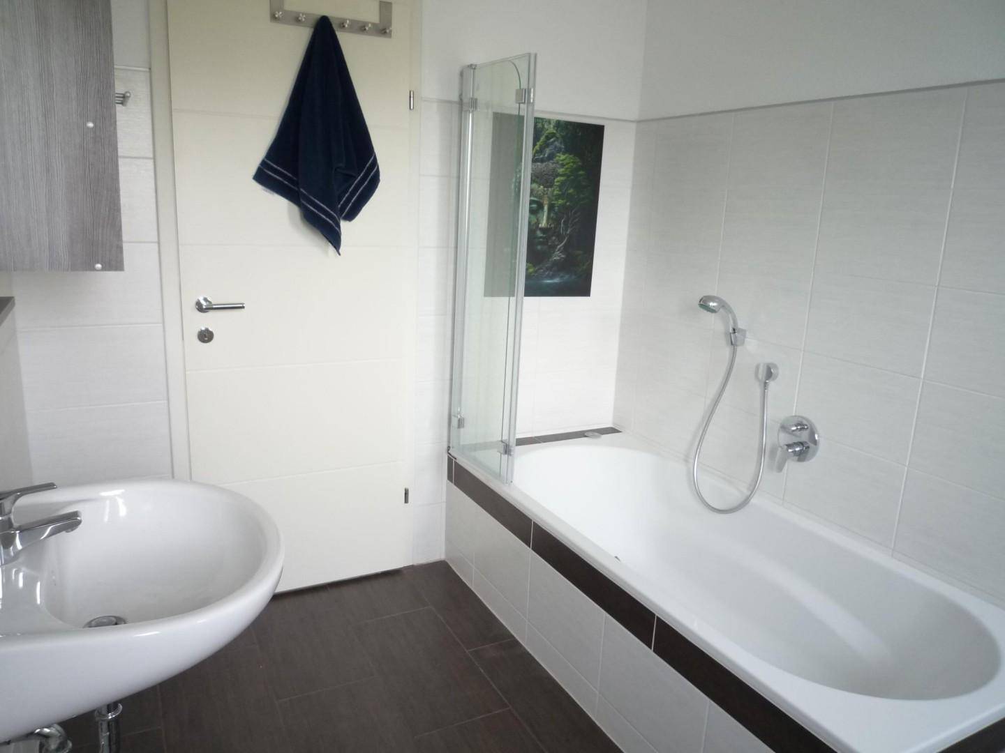 Modernes Badezimmer mit Badewanne, Duschwand, Waschbecken und dunklem Fliesenboden.