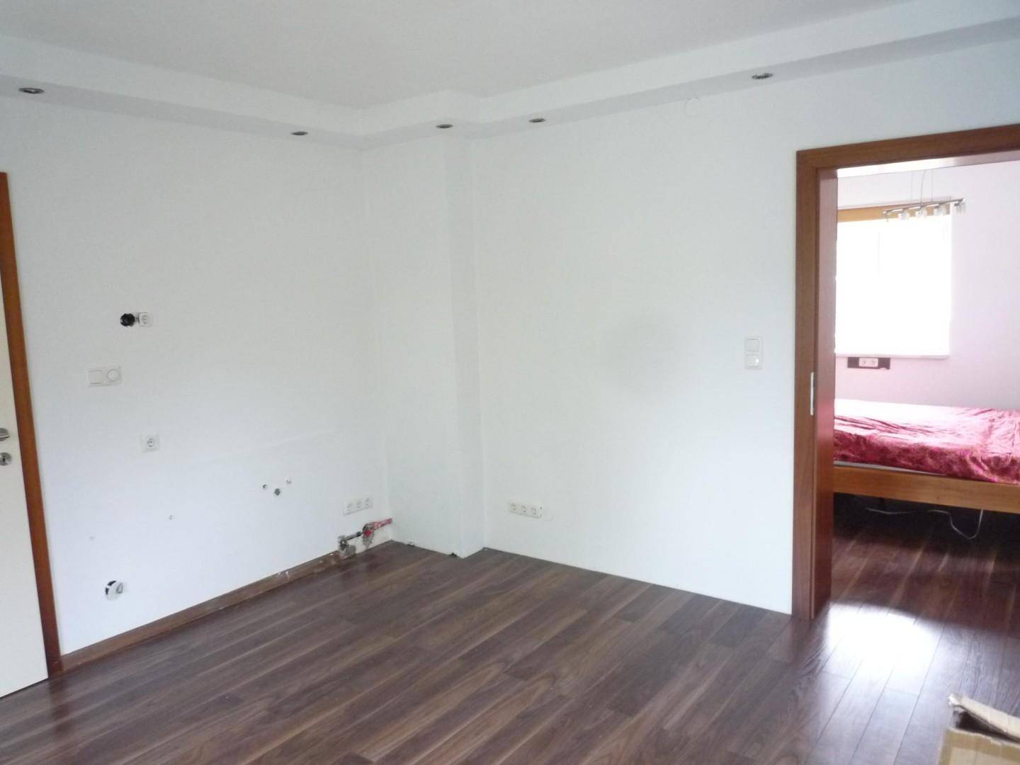 Leerer Raum mit dunklem Laminatboden und weißen Wänden, mit Blick in ein angrenzendes Schlafzimmer.