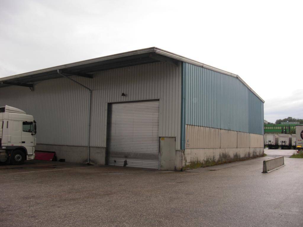 Seitenansicht einer großen Lagerhalle mit Wellblechfassade in Grau und Blau, umgeben von einem nassen Vorplatz.