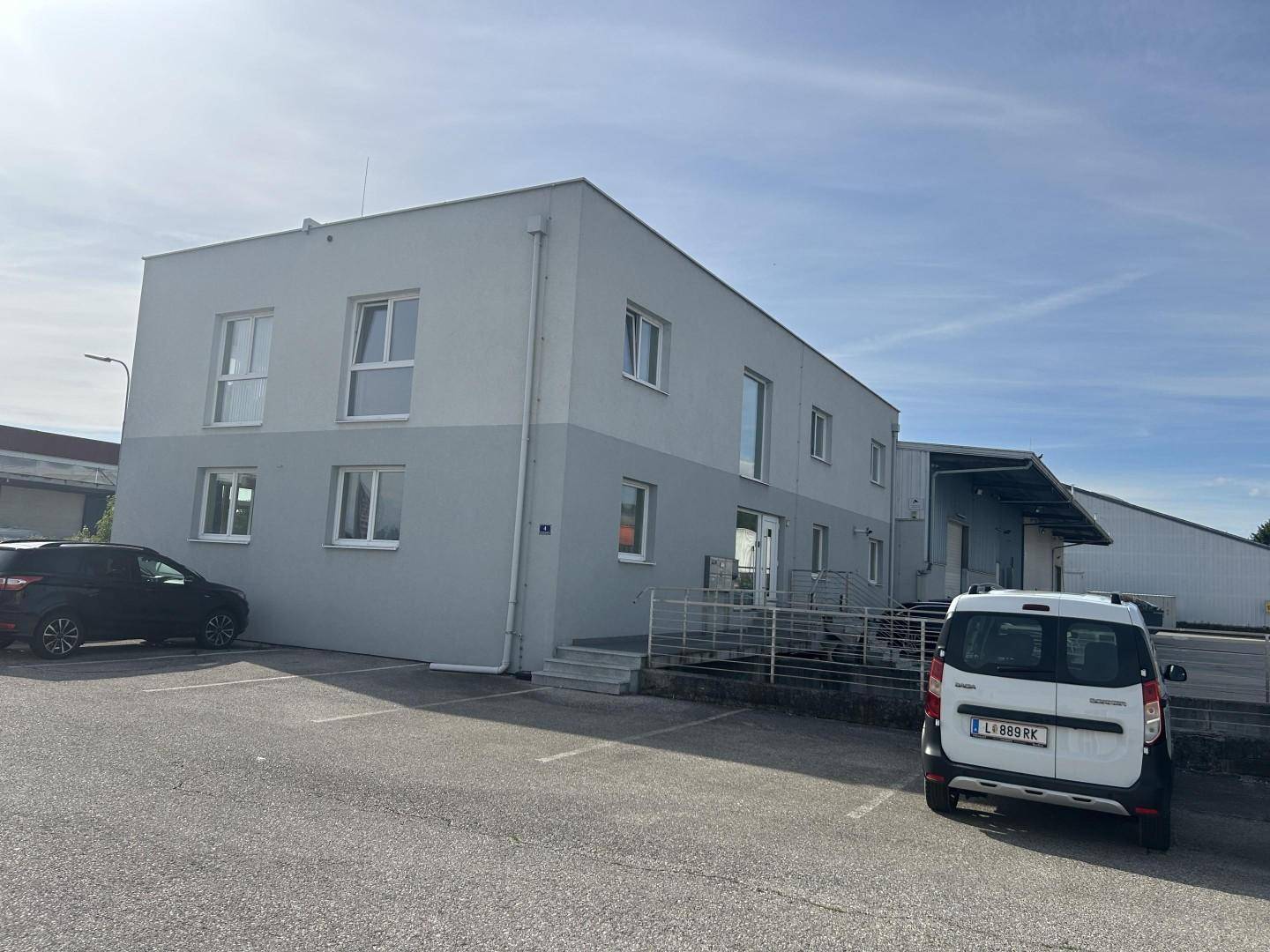 Vorderansicht des modernen Bürogebäudes mit hellgrauer Fassade und mehreren Parkplätzen davor.